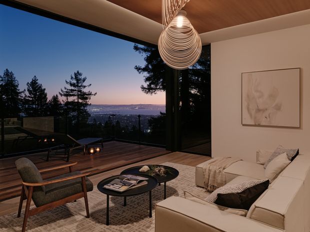 A refined living room opening onto a timber outdoor deck, with expansive floor-to-ceiling glazing overlooking a coastal or waterfront landscape at dusk. The room features a large cream sectional sofa, patterned area rug, mid-century-inspired lounge chair, round dark coffee tables, a timber ceiling, minimalist artwork, and a sculptural woven pendant light. The atmosphere is warm, calm, and inviting.