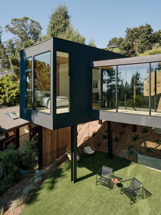 Exterior view of a contemporary elevated addition with a dark, vertically clad box-like volume connected to a glazed pavilion. The building is raised on slender posts above a landscaped sloping garden, with extensive floor-to-ceiling glazing and a small outdoor seating area below. Interior glimpses reveal a bedroom in the enclosed volume and a living area in the glazed section.
