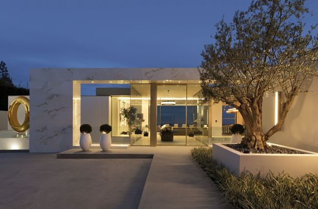 Twilight view of a refined modern residential entrance courtyard. A pale marble-clad rectangular facade frames floor-to-ceiling glazed doors and windows, with warm concealed lighting illuminating the entry. A large sculptural circular feature sits at left, while a mature tree is planted in a raised white rectangular planter with low landscaping at right. The space has clean geometric paving, minimalist detailing, and views through the glazing into a warmly lit interior and beyond.