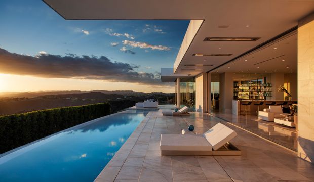 Luxury modern home overlooking a hilly landscape at sunset, with a long infinity-edge swimming pool, expansive outdoor terrace, floor-to-ceiling glazing, and an open-plan interior featuring a kitchen or bar area and extensive shelving. The design uses strong horizontal lines, a projecting flat roof, warm stone finishes, and polished contemporary detailing.