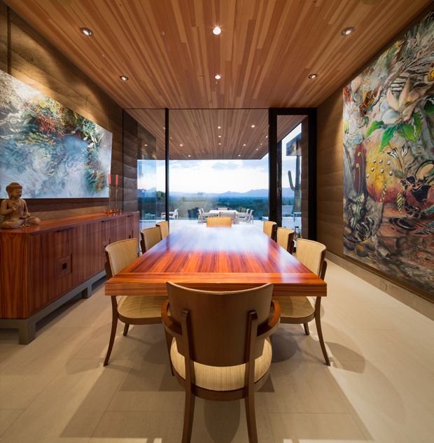 A warm, refined dining room centered on a long polished timber dining table with upholstered timber chairs. The room features extensive timber wall cabinetry, a slatted timber ceiling with recessed lighting, large abstract artworks on both side walls, and full-height glazed doors opening to an outdoor terrace and expansive landscape view.