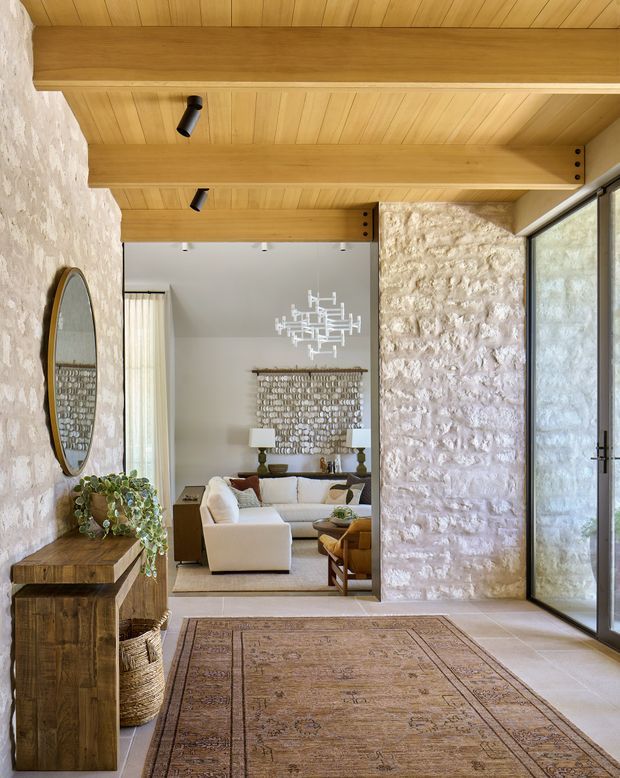 A refined rustic interior view from an entry or hallway into a living room. The space features rough natural stone walls, warm timber ceiling boards and exposed beams, large dark-framed glazed doors, pale stone or tile flooring, and a large patterned rug. A timber console, woven basket, mirror, sofa, sculptural chandelier, and textured wall art are visible, creating a warm, natural, inviting atmosphere.