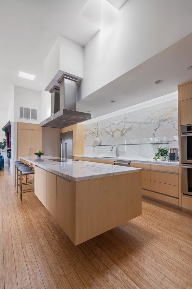 Bright contemporary kitchen with a large central island, pale timber cabinetry, a white veined stone benchtop and splashback, integrated appliances, timber flooring, a stainless-steel rangehood, and a high angled ceiling with skylight-like openings. Bar stools line the island, while a glass shelf and minimal decorative greenery add subtle detail.