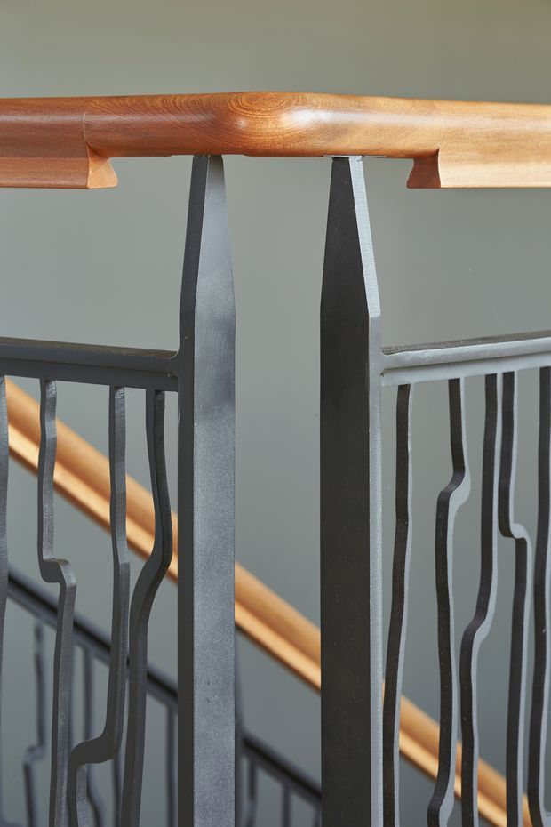 Close-up architectural detail of a stair balustrade, featuring dark grey metal posts and organically shaped vertical balusters beneath a rounded, warm timber handrail. A second timber handrail and railing are visible in the background against a muted grey wall.