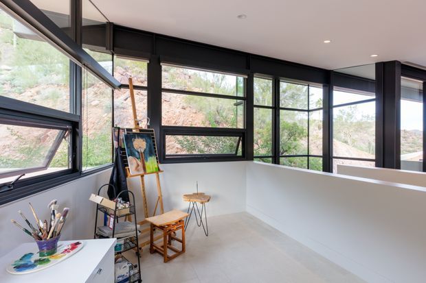 Bright contemporary studio or home office with expansive black-framed windows, a white half-height partition, pale tiled floor, and an easel with art supplies and artwork. The room overlooks a rocky, vegetated hillside and has a clean, airy workspace atmosphere.