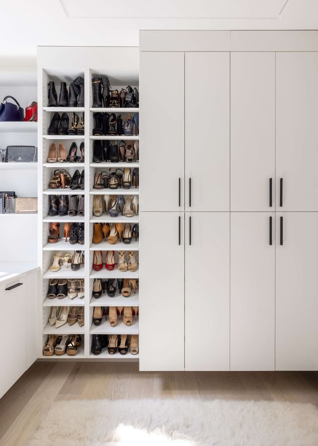 Bright, highly organised walk-in wardrobe with extensive open shelving displaying shoes and handbags alongside tall white built-in cabinets with slim black handles. The room has white walls and cabinetry, a light timber floor, and a soft white rug, creating a clean, refined dressing-room interior.