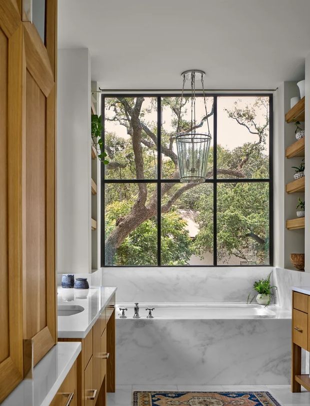A bright, refined bathroom centred on a built-in bathtub beneath a large black-framed, multi-pane window overlooking leafy trees. The room features white marble-look wall and bath surrounds, white surfaces, warm timber cabinetry and shelving, a glass pendant light, and a patterned rug.