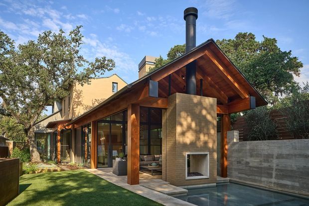 Exterior view of a contemporary single-storey pavilion-style residence with a steep gabled timber roof and exposed timber soffit and beams. Floor-to-ceiling glazing opens toward a landscaped lawn and narrow pool, while a large masonry outdoor fireplace projects from the facade. A darker multi-storey house volume is visible behind the pavilion.