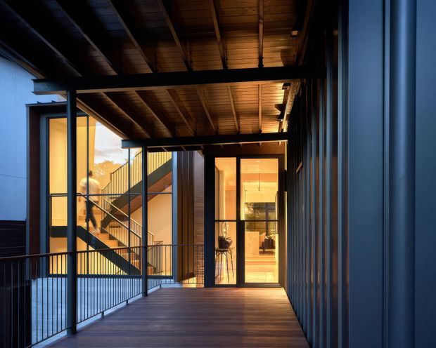 A covered exterior walkway or balcony connecting dark-clad building volumes, with warm timber soffits and decking, black metal framing and balustrades, and large glazed openings revealing a warmly lit interior and stairway beyond. The image has a contemporary architectural character with strong linear structure and a dramatic contrast between cool exterior tones and warm interior lighting.