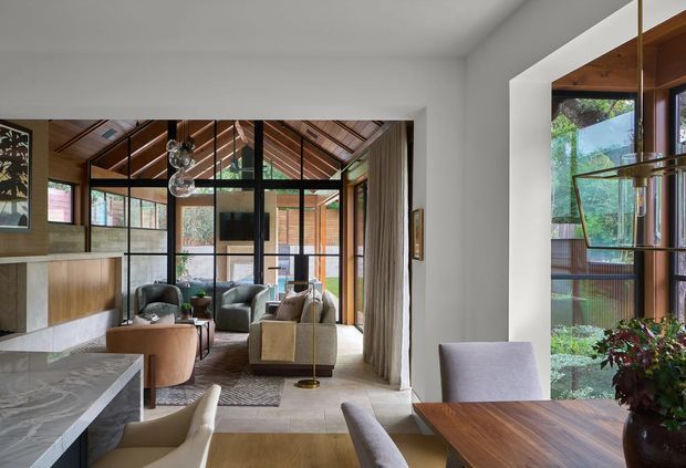 A refined open-plan interior combining a dining area in the foreground with a living room beyond. The space features a dramatic pitched timber-lined ceiling with exposed rafters, extensive black-framed glazing, warm timber joinery, a stone kitchen island, neutral upholstered seating, and a sculptural pendant light. A garden and timber exterior structure are visible through the glazing.