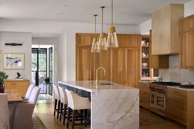 A warm, refined open-plan kitchen and dining space with extensive custom timber cabinetry, a large marble-look island with integrated sink, and seating for several people. The kitchen includes a substantial freestanding range cooker, brass-toned hardware and tapware, and a grey stone backsplash. A dining table and chairs sit to the left, while pendant lights hang above the island. Glazed doors bring natural light into the room.