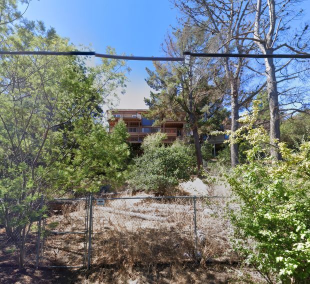 A partially obscured multi-level residential building is viewed through dense trees and shrubs. The house has dark timber or wood-toned balcony railings and cladding, broad outdoor balconies, and a low-pitched roof, set on a dry, wooded site behind a wire fence. Clear blue sky and mature vegetation surround the dwelling.