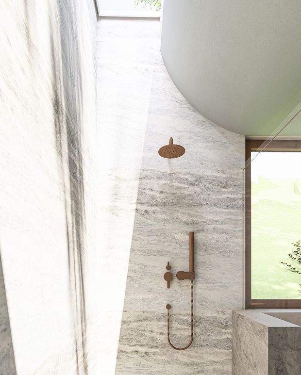 A bright, sculptural bathroom shower area with a curved ceiling, full-height grey-white veined stone wall finish, and a minimalist brushed bronze shower set including an overhead rain shower and handheld shower. A large timber-framed window brings in daylight beside a substantial stone-clad ledge or tub surround.