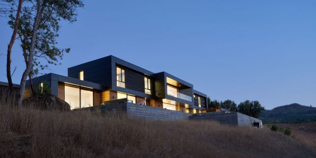A modern multi-level residence viewed from a sloping rural site at dusk. The building has a strongly rectilinear composition with flat roof planes, dark horizontal cladding, expansive illuminated glazing, warm timber-toned sections, and concrete retaining walls. Dry grasses and mature trees frame the foreground, with wooded hills visible in the distance.
