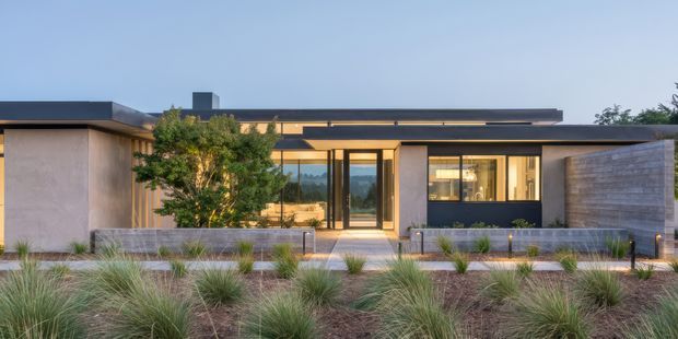 A low-slung single-storey contemporary residence with a strongly horizontal composition, flat rooflines, broad glazing, and muted rendered and concrete-toned exterior walls. A central glazed entry is framed by projecting roof planes, while warm interior lighting is visible through the windows. The building is set behind a landscaped frontage with ornamental grasses and a low masonry boundary wall.