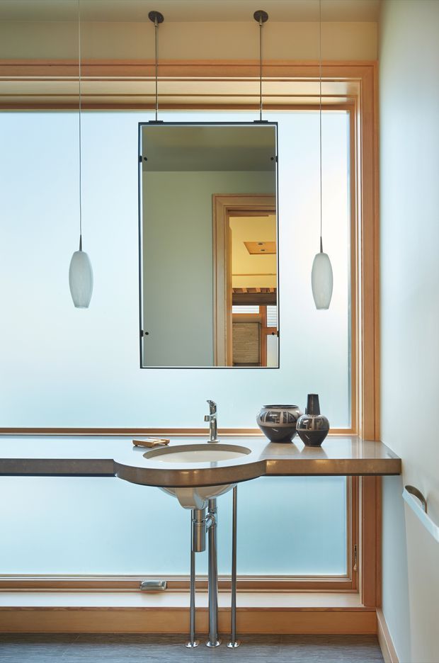 A compact, refined bathroom vanity positioned in front of a large frosted-glass window with timber framing. The floating dark timber-toned benchtop has an integrated oval basin and exposed chrome plumbing, accompanied by a rectangular suspended mirror and two minimalist pendant lights. Timber trim, pale blue-white glazing, and decorative ceramic vessels create a warm modern interior.