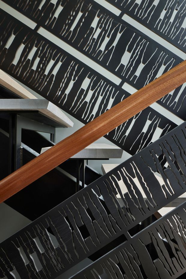 A dramatic interior view of a contemporary multi-level stairway or landing, featuring overlapping black metal stair balustrades or screens with irregular vertical cut-outs. Warm timber handrails cross the composition, while pale concrete or plaster stair elements and dark structural framing create a strong geometric, high-contrast architectural scene.