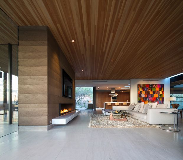 Open-plan contemporary living room with a large white sectional sofa, shag rug and low coffee table, centred around a tall stone-clad fireplace and television wall. The room features extensive warm timber lining across the ceiling, pale large-format flooring, recessed downlights, broad glazing and a kitchen/dining area visible beyond.