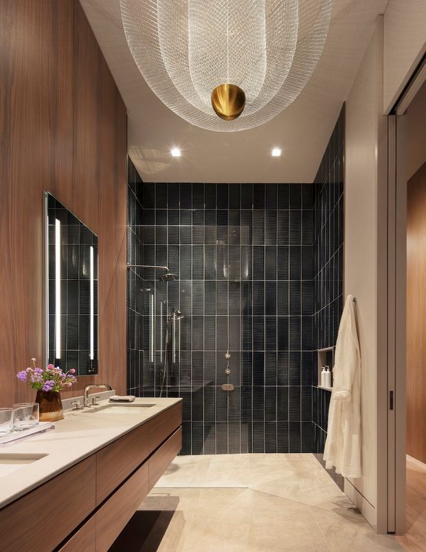 A refined contemporary bathroom with a long floating timber vanity, pale stone benchtop and integrated basin, large illuminated mirror, and a walk-in shower enclosed by clear glass. The shower walls are clad in dark vertically ribbed rectangular tiles, contrasting with warm wood wall panelling and pale stone-look floor tiles. A sculptural woven pendant light with a brass-gold centre hangs from the ceiling, adding a luxurious focal point.