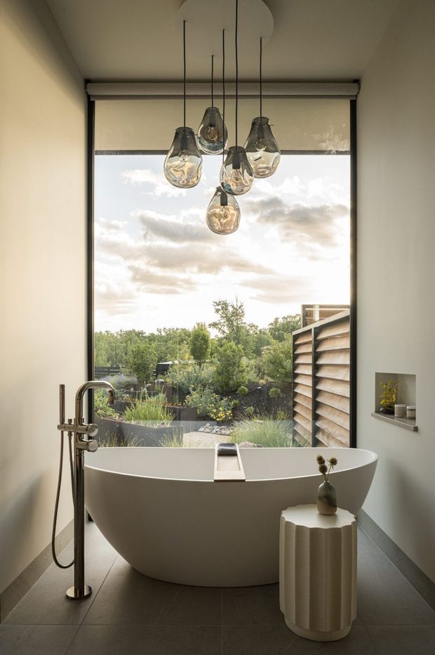 A refined bathroom featuring a freestanding white oval bathtub positioned in front of a large picture window overlooking a landscaped garden. A dark brushed-metal floor-mounted bath mixer, sculptural glass pendant cluster, roller blind, grey floor tiles, and a small fluted pedestal with a vase complete the calm, contemporary space.