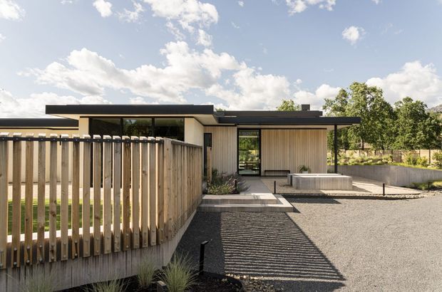 A contemporary single-storey home viewed from the exterior, with a low flat roof, dark horizontal roof edges, pale vertical timber cladding, extensive glazing, and a gravel forecourt. A timber slat fence and concrete pathway lead toward the recessed entry, while established trees and a bright sky form the background.