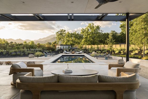 A refined outdoor entertaining area beneath a broad covered pavilion, furnished with a large timber-framed sofa, lounge chairs and a low oval coffee table. The space opens toward a rectangular swimming pool, paved terrace and landscaped garden, with layered trees and distant hills beyond. A dark exposed steel roof structure and slim columns contrast with the pale ceiling and light stone paving, creating a calm indoor-outdoor living setting.