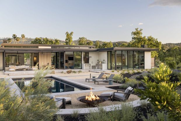 A low, single-storey contemporary home with broad flat rooflines and extensive floor-to-ceiling glazing, viewed from a landscaped courtyard and pool area. The residence opens onto generous paved terraces with outdoor lounge seating, a rectangular swimming pool, and a central outdoor fire pit. The surrounding planting is naturalistic and drought-tolerant, with warm neutral architectural finishes and dark-framed glazing.