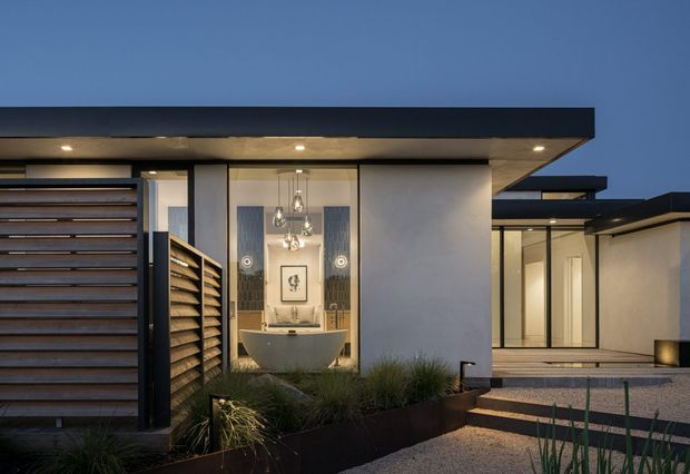 Twilight exterior view of a low-slung contemporary residence. The composition focuses on a covered entry with a dark horizontal roof slab, timber-toned horizontal garage or privacy screening, a glazed entry alcove containing a pedestal basin and pendant lights, and a textured pale rendered wall. Layered flat roof forms and floor-to-ceiling glazing extend toward the right, with restrained architectural planting and gravel paving in the foreground.