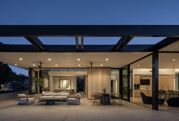 A sophisticated open-plan indoor-outdoor living and dining area photographed at dusk. The space features a broad covered terrace with a dark exposed steel pergola structure, recessed ceiling lights, ceiling fans, expansive black-framed glazing, a low modular sofa arrangement, and a dining table beside a timber-lined central partition. The interior beyond includes a contemporary kitchen and additional seating, creating a seamless connection between the terrace and living spaces.