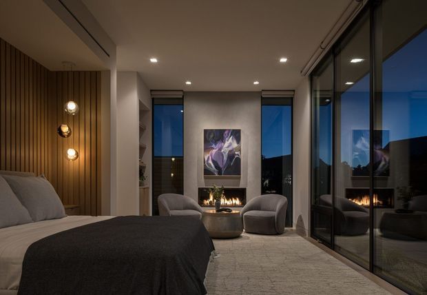 A refined contemporary bedroom at dusk, featuring a low-profile bed with layered neutral bedding, a dark charcoal throw, a vertical timber-slat feature wall with globe pendant lights, and a central fireplace beneath abstract artwork. Floor-to-ceiling black-framed glazing reflects the fireplace and opens views to the dark exterior. Two upholstered lounge chairs and a round coffee table create a small sitting area, while recessed ceiling lighting provides warm ambient illumination.