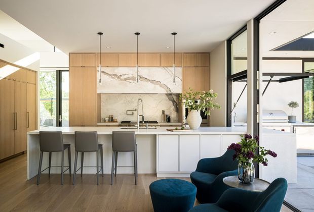 A bright, open-plan kitchen and living area with a large central island, three bar stools, warm timber cabinetry, pale stone benchtops, and a dramatic veined stone splashback. Black-framed floor-to-ceiling glazing connects the interior to an outdoor entertaining area. A deep teal lounge chair and ottoman create a small seating zone in the foreground, with pendant lights suspended above the island.