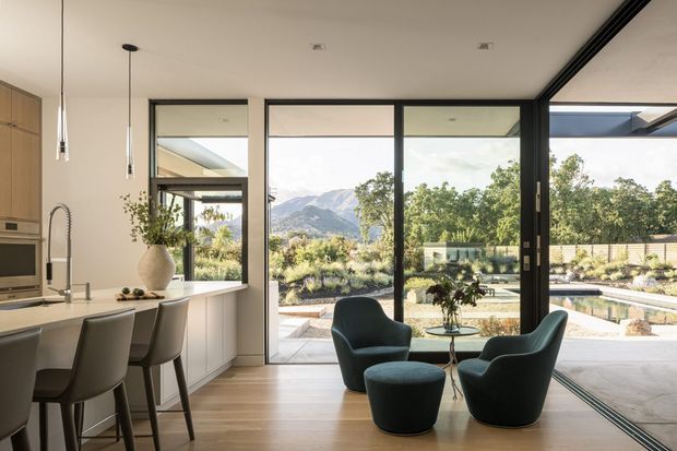 A refined open-plan kitchen and living area with a compact island and integrated seating in the foreground. Full-height black-framed glazing and sliding doors open to a landscaped outdoor area and swimming pool, with mountain views beyond. The interior uses pale timber flooring, white cabinetry and walls, dark-framed windows, sculptural upholstered lounge chairs, and pendant lighting.