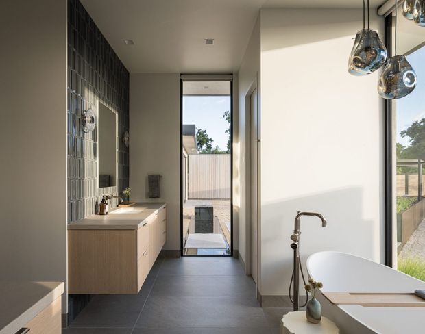 A contemporary bathroom viewed from the entry, featuring a freestanding white bathtub beside large windows, a wall-mounted brushed-metal bath filler, dark grey tiled flooring, a light timber vanity with drawers, and a textured glass-block feature wall. Sculptural pendant lights hang above the bath, while a glazed door opens toward an outdoor courtyard.