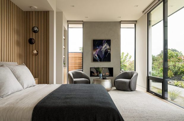 A refined contemporary bedroom with a large upholstered bed in the foreground, a charcoal throw, and a warm vertical timber feature wall behind the headboard. The room includes a small sitting area with two rounded armchairs and a sculptural side table, a fireplace or dark built-in feature beneath abstract artwork, pale carpeting, recessed ceiling lights, and expansive black-framed glazing opening toward a landscaped exterior.