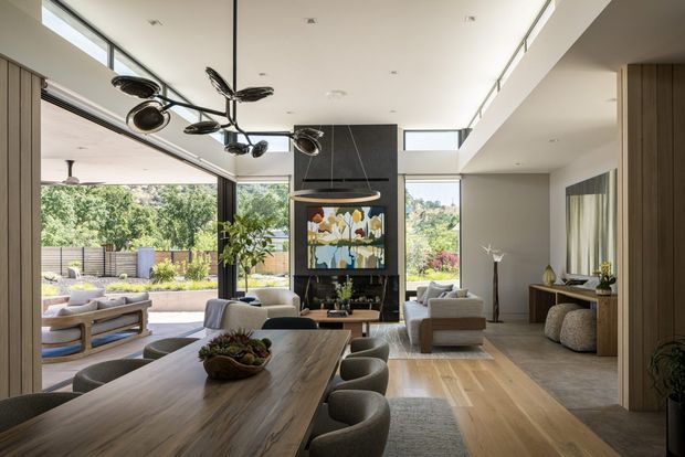 A spacious, refined open-plan living and dining area with floor-to-ceiling glazing opening onto an outdoor terrace and garden. The room features a long timber dining table with upholstered chairs, a contemporary lounge arrangement, a fireplace wall with artwork, light timber flooring, recessed lighting, and a sculptural black pendant light. A timber-clad vertical feature and console area add warmth to the otherwise neutral interior.