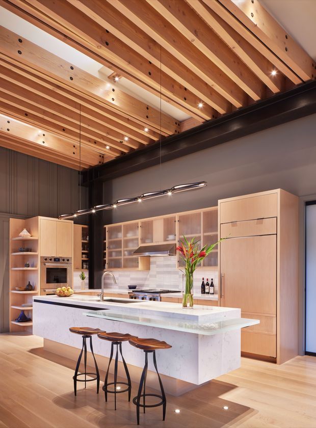 A spacious contemporary kitchen with a large white stone island and integrated sink, three dark-framed timber bar stools, pale timber cabinetry, open shelving, a tiled backsplash, and built-in appliances. The room is defined by exposed timber ceiling beams, dark structural steel framing, recessed and linear pendant lighting, and warm timber flooring.