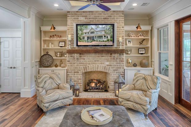 A refined living room centred on a pale brick fireplace with a wood mantel and built-in display shelving. Two upholstered armchairs face a tufted ottoman, with warm timber flooring, white painted wall panelling and trim, a ceiling fan, recessed lighting, and glazed doors and windows. The room has a symmetrical, traditional coastal-country feel.