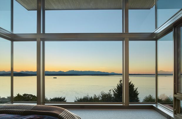 Interior view through expansive floor-to-ceiling corner glazing, overlooking a calm body of water and distant mountain ranges at sunset. Dark metal window frames, a timber-lined ceiling, and a low upholstered seating edge are visible, creating a serene, outlook-focused space.
