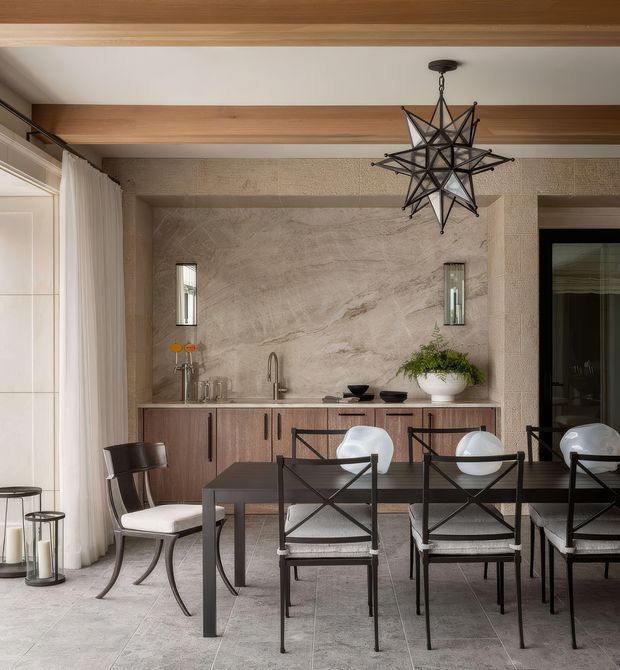 A refined dining area with a long dark dining table and black-framed chairs arranged in front of a built-in buffet and bar cabinet. The room features warm timber ceiling beams, a large textured stone-look feature wall with integrated narrow mirrors, pale tiled flooring, sheer floor-to-ceiling curtains, and a dramatic black geometric star pendant light. A sculptural white vessel with greenery and understated tableware add restrained decorative touches.