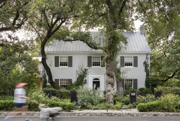 Front exterior view of a two-storey light-painted brick or rendered house, framed by mature trees and dense landscaped planting. The home has a steep standing-seam metal gable roof, symmetrical multi-pane windows with dark shutters, a central entry door with a small pediment, and a low black metal fence along the street.