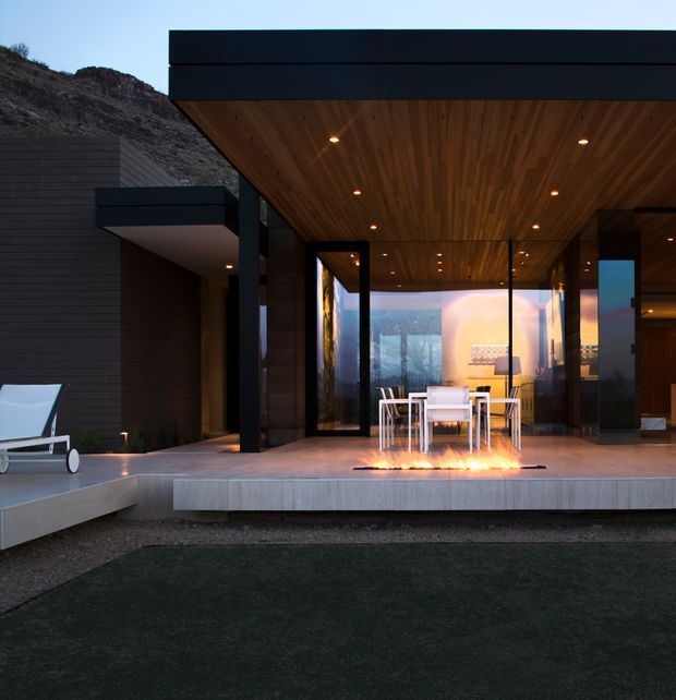 A modern hillside residence viewed from an outdoor terrace. The home features a strong black flat-roofed form, extensive floor-to-ceiling glazing, dark horizontal cladding, and a warm timber-lined soffit with recessed lighting. A dining setting sits beneath the covered terrace beside a linear outdoor fire feature, with a minimalist pool or dark reflecting surface in the foreground and rocky terrain behind.