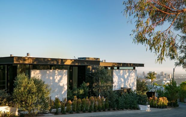 Exterior view of a low, flat-roofed modern home on a landscaped hillside site. The building has dark cladding and framing, large glazed openings, crisp white wall planes, and a projecting horizontal roofline. Drought-tolerant planting surrounds the house, with a distant urban skyline visible beyond.