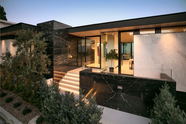 A contemporary single-storey house facade at dusk, featuring a low flat roof, extensive floor-to-ceiling glazing, black marble-look wall surfaces, pale stone or plaster wall panels, and a raised entry reached by illuminated steps. The landscaped foreground includes drought-tolerant planting, while warm interior lighting reveals an entry and living area behind the glazing.