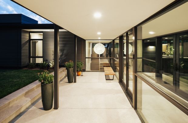 A covered, contemporary courtyard passage connecting dark-clad building volumes. The space has a smooth pale floor, a flat illuminated ceiling, extensive black-framed glazing on the right, dark horizontal wall cladding on the left, built-in bench seating, and potted plants. A small landscaped courtyard and garden are visible to the left, while interior spaces are visible through the glazing.
