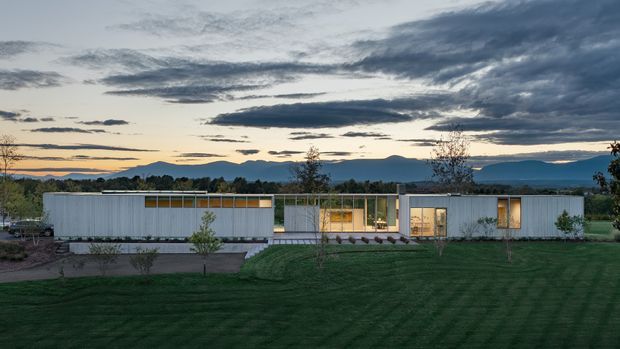 A long, single-storey contemporary residence set in a landscaped suburban or rural site, photographed at dusk. The building has a low horizontal profile, pale vertically ribbed cladding, extensive floor-to-ceiling glazing, and warm interior lighting. A broad lawn and young trees sit in the foreground, with distant hills and a dramatic cloudy sky beyond.