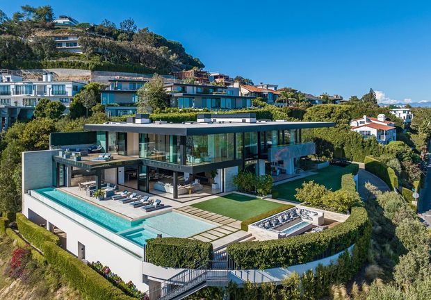 A large contemporary multi-level hillside residence with extensive floor-to-ceiling glazing, broad flat roof planes, terraces, landscaped gardens, and an outdoor swimming pool. The property overlooks a suburban coastal-looking setting, with additional hillside homes visible in the background. A separate spa or water feature sits beside the pool, and the house uses a strong indoor-outdoor layout with expansive entertainment areas.