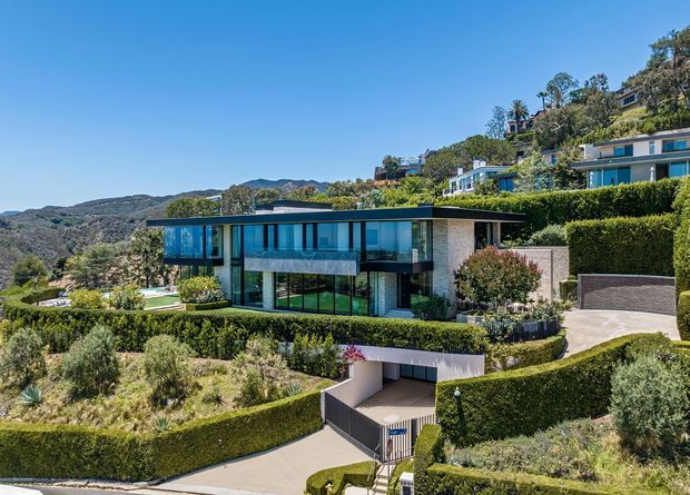 Wide exterior view of a contemporary multi-level residence set on a steep, landscaped hillside. The home features extensive floor-to-ceiling glazing, dark structural framing, pale masonry walls, flat rooflines, terraces, manicured hedges, and a driveway leading to an integrated lower-level garage. Other hillside homes and wooded terrain are visible in the background.