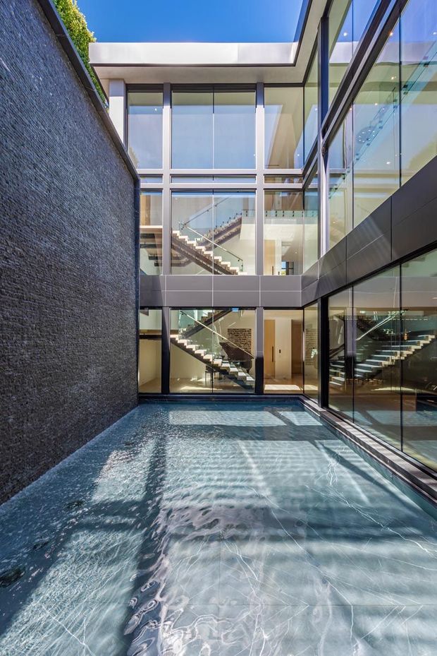 A bright contemporary multi-level courtyard viewed from a narrow outdoor pool. Dark textured masonry forms one side, while extensive floor-to-ceiling glazing reveals internal staircases and a double-height circulation space. The building has crisp rectangular forms, dark metal framing, a flat roof opening to the sky, and strong reflections across the water and glass.