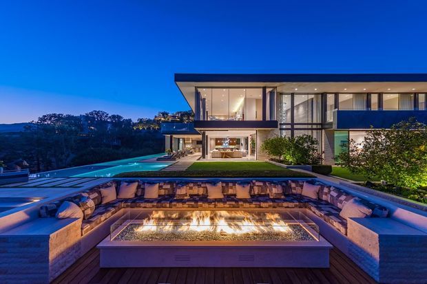 Twilight exterior view of a large contemporary multi-storey home with extensive floor-to-ceiling glazing, projecting flat roof planes, dark horizontal facade bands, and landscaped grounds. A long swimming pool and a sunken outdoor lounge with built-in seating surround a linear outdoor fire feature in the foreground.