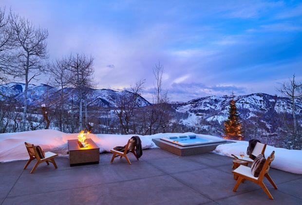 A spacious outdoor terrace overlooking snow-covered mountains at dusk. The terrace has concrete paving, several timber lounge chairs, a square outdoor fire pit with flames, and a raised rectangular spa or plunge pool. Snowbanks line the terrace, with a lit evergreen tree and leafless trees framing the mountain landscape. The scene has a cool blue winter atmosphere with warm firelight accents.